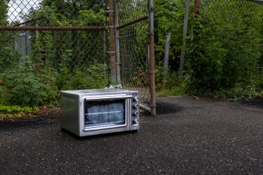 A discarded toaster oven sits abandoned on a wet pavement in front of a chain-link fence. The appliance shows signs of neglect, blending into the overgrown urban surroundings. clipart