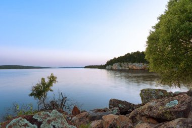 Atoka Oklahoma Parkı 'nda sakin göl sularının gökyüzünün yumuşak renklerini yansıttığı sakin bir alacakaranlık sahnesi. Kayalık kıyı şeridi ve sık ağaç şeridi doğa severler için mükemmel huzurlu ve manzaralı bir ortam yaratır..