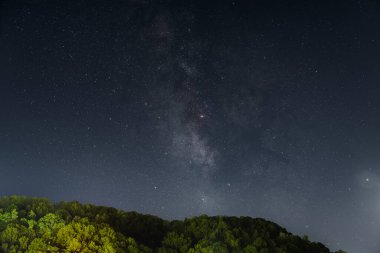 Samanyolu 'nun nefes kesen manzarası ve ağaçlarla kaplı bir tepenin üstündeki gece gökyüzünde sayısız yıldız. Karanlık gökyüzü, yıldızları seyretmek ve astrofotoğrafçılık için mükemmel çarpıcı bir gökyüzü yaratır..