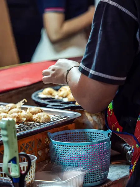 Tayland 'da bir sokak yemeği satıcısı küçük bir ızgarada abur cubur pişirir. Önlük ve mavi gömlek giyerek, yakınlarda mavi bir sepeti olan yiyeceklere dikkatle bakarlar.