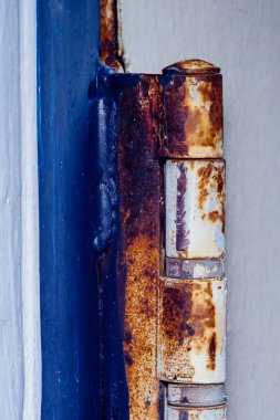 Paslı bir menteşeye yakın çekim, belirgin bozulma ve aşınma gösteriyor, canlı mavi bir kapıya bağlı, yıpranmanın ve zamanın etkilerini vurguluyor.