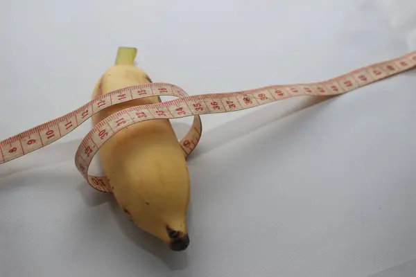 Ölçüm bandıyla sarılı kısa sarı bir muz, temiz beyaz bir arkaplan üzerine yerleştirilmiş. Bu resim, ölçüm, sağlık ve gıda porsiyonları ile ilgili kavramlara uygun olarak, meyvenin uzunluğunu ve çevresini vurgular..