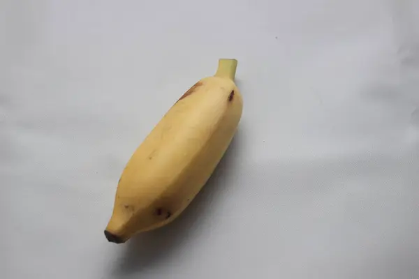 Beyaz desenli arka plana yerleştirilmiş tek bir olgun sarı muz. Meyve, hafif kahverengi benekli, doğal ışıklandırmayla fotoğraflanmış, yiyecek, beslenme veya tropikal meyvelerle ilgili kavramlar için idealdir..