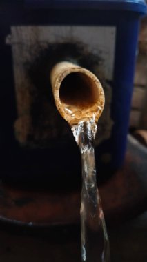 Temiz su, açık kahverengi bir borunun ucundan sürekli akar ve arka planda bulanık bir konteynır.