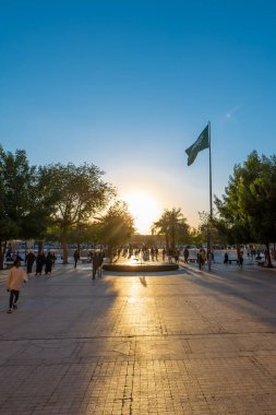 Arka planda Suudi bayrağı olan bir Riyad Çeşmesi Günbatımı Manzarası. Çeşme, Suudi Arabistan 'ın Riyad ilinin Ad Dirah ilçesinde Al-Safat Meydanı yakınlarında yer almaktadır. Günbatımı Suudi bayrağı için güzel bir zemin sağlar..