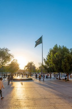 Arka planda Suudi bayrağı olan bir Riyad Çeşmesi Günbatımı Manzarası. Çeşme, Suudi Arabistan 'ın Riyad ilinin Ad Dirah ilçesinde Al-Safat Meydanı yakınlarında yer almaktadır. Günbatımı Suudi bayrağı için güzel bir zemin sağlar..