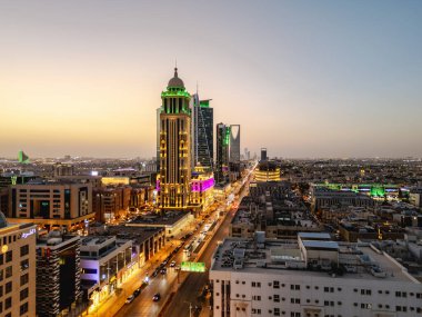 Aydınlatılmış Riyad şehrinin hava aracı görüntüsü, önde gelen kuleler ve çağdaş kentsel tasarım da dahil olmak üzere modern gökyüzünü gösteriyor.