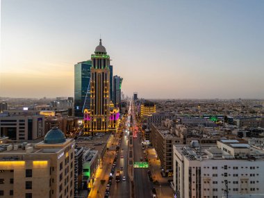 Aydınlatılmış Riyad şehrinin hava aracı görüntüsü, önde gelen kuleler ve çağdaş kentsel tasarım da dahil olmak üzere modern gökyüzünü gösteriyor.