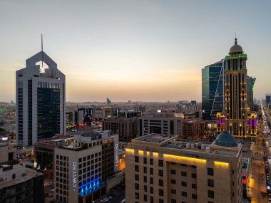 Aydınlatılmış Riyad şehrinin hava aracı görüntüsü, önde gelen kuleler ve çağdaş kentsel tasarım da dahil olmak üzere modern gökyüzünü gösteriyor.