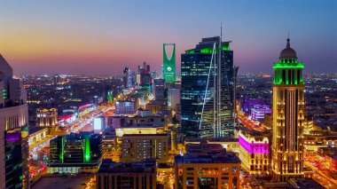 Aydınlatılmış Riyad şehrinin hava aracı görüntüsü, önde gelen kuleler ve çağdaş kentsel tasarım da dahil olmak üzere modern gökyüzünü gösteriyor.