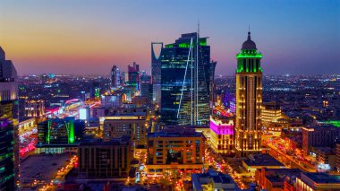 Aydınlatılmış Riyad şehrinin hava aracı görüntüsü, önde gelen kuleler ve çağdaş kentsel tasarım da dahil olmak üzere modern gökyüzünü gösteriyor.