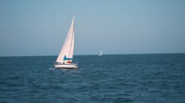 Gün batımında sakin bir denizde süzülen bir yelkenlinin yavaş çekim, sinematik çekimi. Yelkenli, uzun direği ve parlak beyaz gövdesiyle sakin sulara karşı durmaktadır. Ufuk çizgisi.