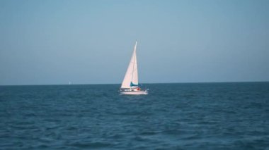 Gün batımında sakin bir denizde süzülen bir yelkenlinin yavaş çekim, sinematik çekimi. Yelkenli, uzun direği ve parlak beyaz gövdesiyle sakin sulara karşı durmaktadır. Ufuk çizgisi.