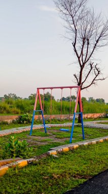  Görüntü, park ortamındaki tek bir salıncağı yakalıyor. Salıncak metalden yapılmıştır ve canlı pembe ve mavi renk düzeniyle boyanmıştır. Beton bir yol ve küçük, çıplak bir ağaçla çevrili yeşil bir çimlerin üzerinde duruyor..