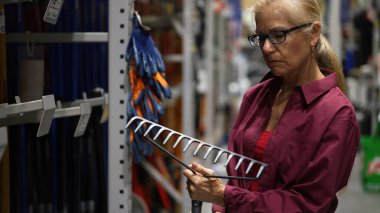 Çekici, mutlu, olgun bir kadın bahçe aletlerine bakıyor ve onu büyük bir hırdavatçıda yakından inceliyor..