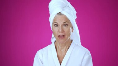 Banyodan çıkmış kafasında havlu olan olgun bir kadının portresi şaşırmış, şaşırmış ve 50 'li yaşlarda yaşlı bir kadının yüzüne dokunmuş, pembe arka plan stüdyosunda yalnız poz vermiş..