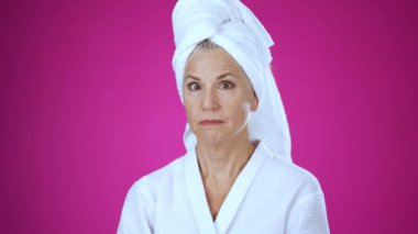 Banyodan çıkmış kafasında havlu olan olgun bir kadının portresi şaşırmış, şaşırmış ve 50 'li yaşlarda yaşlı bir kadının yüzüne dokunmuş, pembe arka plan stüdyosunda yalnız poz vermiş..