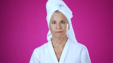 Banyodan çıkmış kafasında havlu olan olgun bir kadının portresi şaşırmış, şaşırmış ve 50 'li yaşlarda yaşlı bir kadının yüzüne dokunmuş, pembe arka plan stüdyosunda yalnız poz vermiş..