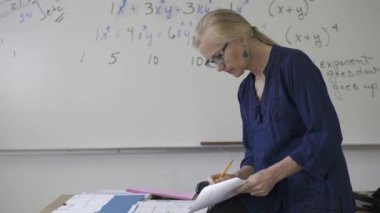 Lise matematik öğretmeni masada oturmuş öğrenciler için ödevler hazırlıyor. Yukarı bakıyor ve gülümsüyor..