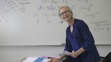 Lise matematik öğretmeni masada oturmuş öğrenciler için ödevler hazırlıyor. Yukarı bakıyor ve gülümsüyor..