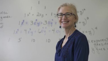 Lise matematik öğretmeni masada oturmuş öğrenciler için ödevler hazırlıyor. Yukarı bakıyor ve gülümsüyor..