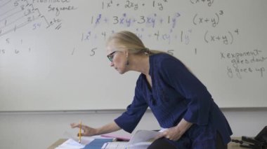 Lise matematik öğretmeni masada oturmuş öğrenciler için ödevler hazırlıyor. Yukarı bakıyor ve gülümsüyor..