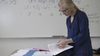 Lise matematik öğretmeni masada oturmuş öğrenciler için ödevler hazırlıyor. Yukarı bakıyor ve gülümsüyor..