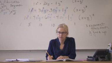 Lise matematik öğretmeni masada oturmuş öğrenciler için ödevler hazırlıyor. Yukarı bakıyor ve gülümsüyor..