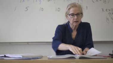 Lise matematik öğretmeni masada oturmuş öğrenciler için ödevler hazırlıyor. Yukarı bakıyor ve gülümsüyor..