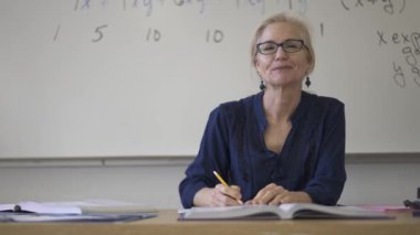 Lise matematik öğretmeni masada oturmuş öğrenciler için ödevler hazırlıyor. Yukarı bakıyor ve gülümsüyor..