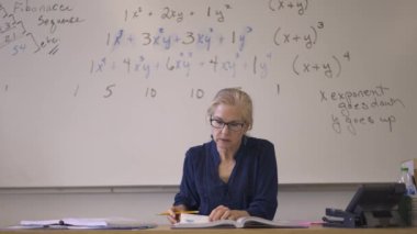 Lise matematik öğretmeni masada oturmuş öğrenciler için ödevler hazırlıyor. Yukarı bakıyor ve gülümsüyor..