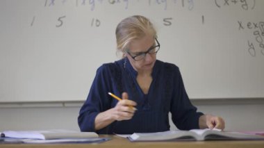 Lise matematik öğretmeni masada oturmuş öğrenciler için ödevler hazırlıyor. Yukarı bakıyor ve gülümsüyor..