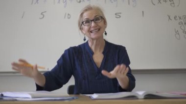 Lise matematik öğretmeni masada oturmuş öğrenciler için ödevler hazırlıyor. Yukarı bakıyor ve gülümsüyor..