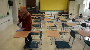 Mutlu Müslüman kadın öğretmen başörtüsü takıyor beyaz tahtaya dünya dinleri yazıyor. Sınıfta Amerikan bayrağı ve arka planda akıllı tahta var..