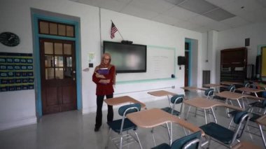 Mutlu Müslüman kadın öğretmen başörtüsü takıyor beyaz tahtaya dünya dinleri yazıyor. Sınıfta Amerikan bayrağı ve arka planda akıllı tahta var..