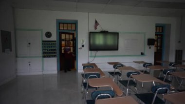 Mutlu Müslüman kadın öğretmen başörtüsü takıyor beyaz tahtaya dünya dinleri yazıyor. Sınıfta Amerikan bayrağı ve arka planda akıllı tahta var..