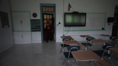 Mutlu Müslüman kadın öğretmen başörtüsü takıyor beyaz tahtaya dünya dinleri yazıyor. Sınıfta Amerikan bayrağı ve arka planda akıllı tahta var..