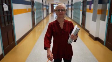 Okulda boş bir koridorda kameraya doğru yürüyen mutlu ve olgun bir kadın öğretmenin yavaş çekimde portresi..