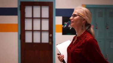 Okulda boş bir koridorda kameraya doğru yürüyen mutlu ve olgun bir kadın öğretmenin yavaş çekimde portresi..