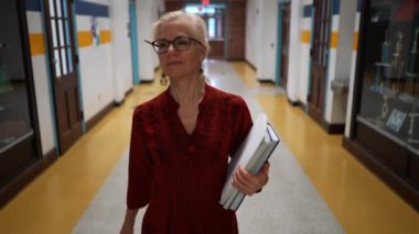 Okulda boş bir koridorda kameraya doğru yürüyen mutlu ve olgun bir kadın öğretmenin yavaş çekimde portresi..