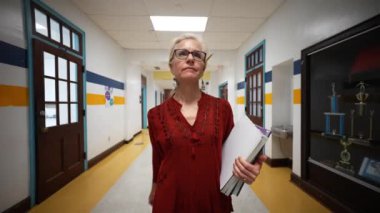 Okulda boş bir koridorda kameraya doğru yürüyen mutlu ve olgun bir kadın öğretmenin yavaş çekimde portresi..