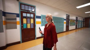 Okulda boş bir koridorda kameraya doğru yürüyen mutlu ve olgun bir kadın öğretmenin yavaş çekimde portresi..