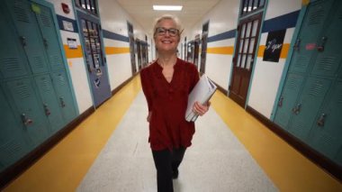 Okulda boş bir koridorda kameraya doğru yürüyen mutlu ve olgun bir kadın öğretmenin yavaş çekimde portresi..