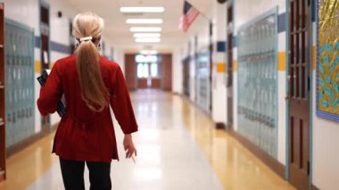 Okulda boş bir koridorda kameraya doğru yürüyen mutlu ve olgun bir kadın öğretmenin yavaş çekimde portresi..