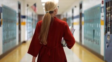 Okulda boş bir koridorda kameraya doğru yürüyen mutlu ve olgun bir kadın öğretmenin yavaş çekimde portresi..
