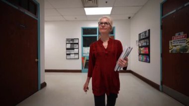 Okulda boş bir koridorda kameraya doğru yürüyen mutlu ve olgun bir kadın öğretmenin yavaş çekimde portresi..