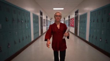 Okulda boş bir koridorda kameraya doğru yürüyen mutlu ve olgun bir kadın öğretmenin yavaş çekimde portresi..