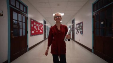 Okulda boş bir koridorda kameraya doğru yürüyen mutlu ve olgun bir kadın öğretmenin yavaş çekimde portresi..