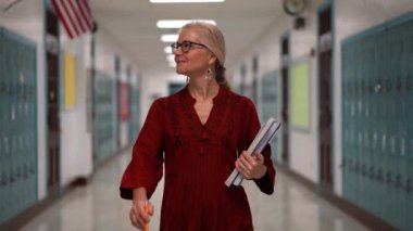 Okulda boş bir koridorda kameraya doğru yürüyen mutlu ve olgun bir kadın öğretmenin yavaş çekimde portresi..