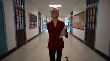 Okulda boş bir koridorda kameraya doğru yürüyen mutlu ve olgun bir kadın öğretmenin yavaş çekimde portresi..